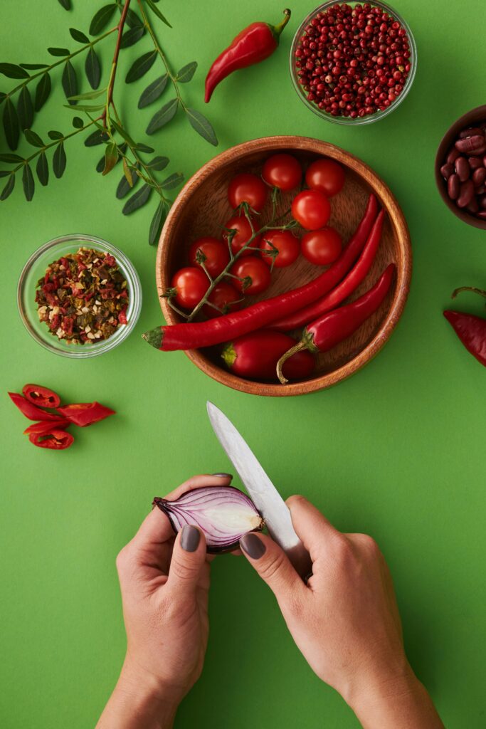 Vibrant top view of fresh vegetables and spices with hands slicing onion on green background.